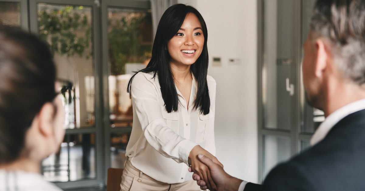 Business, career and placement concept - image from back of two employers sitting in office and shaking hand of young asian woman after successful negotiations or interview